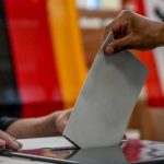 
              A man casts his ballot for the German elections in a polling station in Berlin, Germany, Sunday, Sept. 26, 2021. In background the German national flag. (AP Photo/Michael Probst)
            