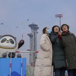 A woman wearing a face mask to help protect from the coronavirus takes a selfie with her companion against a Beijing Winter Olympics Mascot and Olympic Tower in Beijing, Wednesday, Jan. 19, 2022. China has locked down parts of Beijing's Haidian district following the detection of three cases, just weeks before the capital is to host the Winter Olympic Games. (AP Photo/Andy Wong)