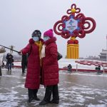 
              FILE - Women wearing face masks printed with a slogan for the Beijing Winter Olympics Games take a selfie with a decoration for the Winter Olympics Games in Tiananmen Square in Beijing on Jan. 20, 2022. The omicron variant is making its latest appearance, and Beijing is locking down some residential communities as the country anxiously awaits the start of the Winter Olympics on Feb. 4. (AP Photo/Andy Wong, File)
            