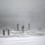 A pedestrian walks through a gust of wind along the waterfront in India Point Park in Providence, R.I., Saturday, Jan. 29, 2022. A powerful nor'easter swept up the East Coast on Saturday, threatening to bury parts of 10 states under deep, furiously falling snow accompanied by coastal flooding and high winds that could cut power and leave people shivering in the cold weather expected to follow. (AP Photo/David Goldman)