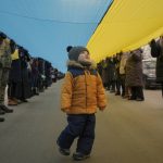 
              A child walks under a large Ukrainian flag carried by people marking a "day of unity" in Sievierodonetsk, the Luhansk region, eastern Ukraine, Wednesday, Feb. 16, 2022. The flags celebrated survival, endurance and above all, defiance. One blue and yellow banner curved along the edge of a stadium field. Others were tiny handheld things. One made it on the Olympic podium. Ukraine's president declared Wednesday a day of national unity in the face of “hybrid threats.” (AP Photo/Vadim Ghirda)
            