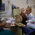 
              Kate Forte kisses her daughter, Lexie Stroiney, 6, as research nurse Michelle Harris, left, demonstrates a blood draw on her stuffed animal "Sprinkles" at Children's National Hospital, in Washington, Wednesday, Jan. 26, 2022. Lexie had COVID-19 and is part of a NIH-funded multi-year study at Children's National Hospital to look at impacts of COVID-19 on children's physical health and quality of life. (AP Photo/Carolyn Kaster)
            