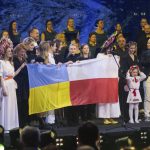 
              Seven-year-old Amellia Anisovych, a refugee from Ukraine, holds her headdress as she stands next to a polish flag in the finale of a fund-raising concert in Lodz, Poland, Sunday, March 20, 2022. Anisovych opened the concert by singing Ukraine's national anthem. She became widely known for singing of a song from the movie Frozen in a bomb shelter in Kyiv in early March. She has since come to Poland with her grandmother and brother. Her parents remained in Kyiv. (AP Photo/Marian Zubrzycki)
            
