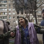 
              People help an elderly woman to walk in a street with an apartment building hit by shelling in the background in Mariupol, Ukraine, Monday, March 7, 2022. (AP Photo/Evgeniy Maloletka)
            