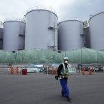 
              A worker helps direct a truck driver as he stands near tanks, background, that will be used to store treated radioactive water after it was used to cool down melted fuel at the Fukushima Daiichi nuclear power plant, run by Tokyo Electric Power Company Holdings (TEPCO), in Okuma town, northeastern Japan, Thursday, March 3, 2022. Treated radioactive water is stored in tanks at the power station. The government has announced plans to release the water after treatment and dilution to well below the legally releasable levels through a planned undersea tunnel at a site about 1 kilometer offshore.  (AP Photo/Hiro Komae)
            