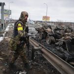 
              An armed man stands by the remains of a Russian military vehicle in Bucha, close to the capital Kyiv, Ukraine, Tuesday, March 1, 2022. Russia on Tuesday stepped up shelling of Kharkiv, Ukraine's second-largest city, pounding civilian targets there. Casualties mounted and reports emerged that more than 70 Ukrainian soldiers were killed after Russian artillery recently hit a military base in Okhtyrka, a city between Kharkiv and Kyiv, the capital. (AP Photo/Serhii Nuzhnenko)
            