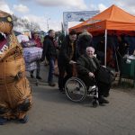 
              A refugee in a wheelchair smiles as a volunteer dressed in a dinosaur suit stands at the border crossing in Medyka, southeastern Poland, Monday, April 11, 2022. (AP Photo/Sergei Grits)
            