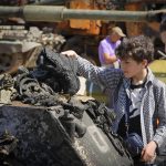 
              A boy holds a boot of a killed Russian soldier at a destroyed Russian tank installed as a symbol of war in central Kyiv, Ukraine, Tuesday, June 7, 2022. St Michael cathedral is in the background. (AP Photo/Efrem Lukatsky)
            