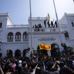 
              A protester, carrying national flag, stands with others on top of the building of Sri Lankan Prime Minister Ranil Wickremesinghe's office, demanding he resign after president Gotabaya Rajapaksa fled the country amid economic crisis in Colombo, Sri Lanka, Wednesday, July 13, 2022. Rajapaksa fled on a military jet on Wednesday after angry protesters seized his home and office, and appointed Prime Minister Ranil Wickremesinghe as acting president while he is overseas. Wickremesinghe quickly declared a nationwide state of emergency to counter swelling protests over the country's economic and political collapse. (AP Photo/Eranga Jayawardena)
            