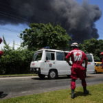 
              members of the Cuban Red Cross standby as a huge plume of smoke rises from the Matanzas Supertanker Base, as firefighters work to quell a blaze which began during a thunderstorm the night before, in Matazanas, Cuba, Saturday, Aug. 6, 2022. Cuban authorities say lightning struck a crude oil storage tank at the base, causing a fire that led to four explosions which injured more than 50 people. (AP Photo/Ramon Espinosa)
            