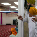 
              Arender Singh, above, places his hand on the shoulder of Amandeep Singh, 12, at the threshold of the prayer room where the Gur Granth Sahib, the Sikh holy book, resides during prayer hours at Guru Nanak Darbar of Long Island, a Sikh gurudwara, Wednesday, Aug. 24, 2022, in Hicksville, N.Y. Their Afghan Sikh family of 13 has found refuge in the diaspora community on Long Island where the Sikh community is helping family members obtain work permits, housing, healthcare and find schools for the children. (AP Photo/John Minchillo)
            