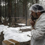 
              Expert smokes under the rain during the exhumation of bodies in the recently retaken area of Izium, Ukraine, Friday, Sept. 16, 2022. Ukrainian authorities discovered a mass burial site near the recaptured city of Izium that contained hundreds of graves. It was not clear who was buried in many of the plots or how all of them died, though witnesses and a Ukrainian investigator said some were shot and others were killed by artillery fire, mines or airstrikes. (AP Photo/Evgeniy Maloletka)
            