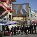
              FILE - Residents walk through a security checkpoint into the Hotan Bazaar where a screen shows Chinese President Xi Jinping in Hotan in western China's Xinjiang region on Nov. 3, 2017. Chinese President Xi Jinping was the son of a communist revolutionary leader, a victim of the Cultural Revolution and a provincial leader who promoted economic growth before ascending to the very top a decade ago. (AP Photo/Ng Han Guan, File)
            