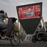 
              FILE - A woman on her electric-powered scooter films a large video screen outside a shopping mall showing Chinese President Xi Jinping speaking during an event to commemorate the 100th anniversary of China's Communist Party at Tiananmen Square in Beijing on July 1, 2021. Chinese President Xi Jinping was the son of a communist revolutionary leader, a victim of the Cultural Revolution and a provincial leader who promoted economic growth before ascending to the very top a decade ago. (AP Photo/Andy Wong, File)
            