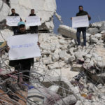 
              FILE - Men stand amidst the rubble of destroyed houses with placards criticizing the United Nations, in Atareb, Syria, Sunday, Feb. 12, 2023. The massive earthquake that hit last week is the latest in a litany of hardships for Syrian women, many of whom have been left dependent on aid and alone responsible for their families' well-being. (AP Photo/Hussein Malla, File)
            