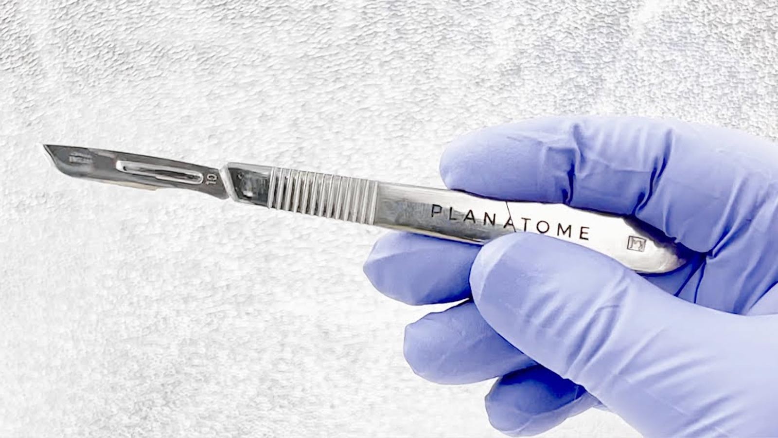 A gloved hand holds a Planatome surgical blade...