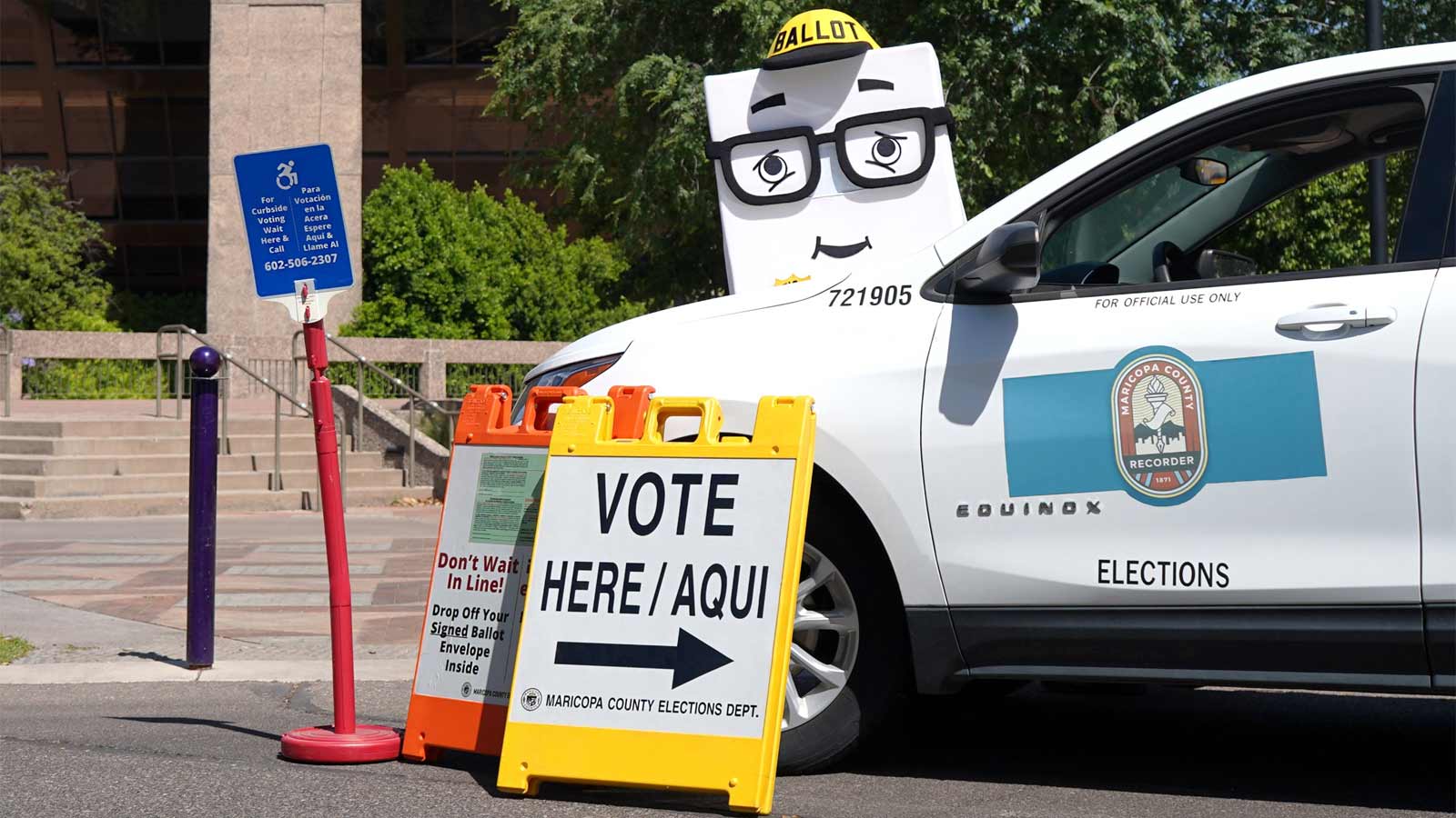 Maricopa County Elections Department mascot Phil the Ballot stands behind a county car and a sign t...