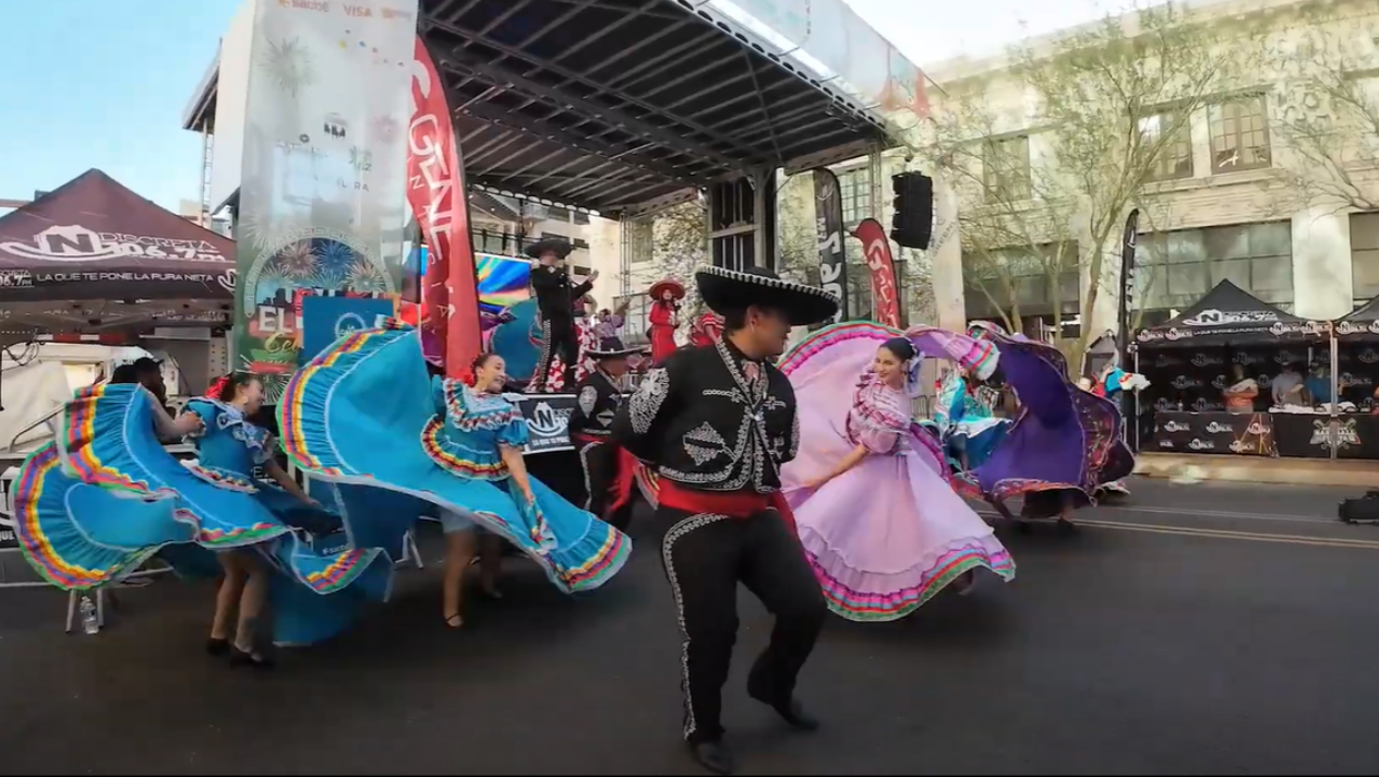 A parade of people in bright dresses and black mariachi suit....