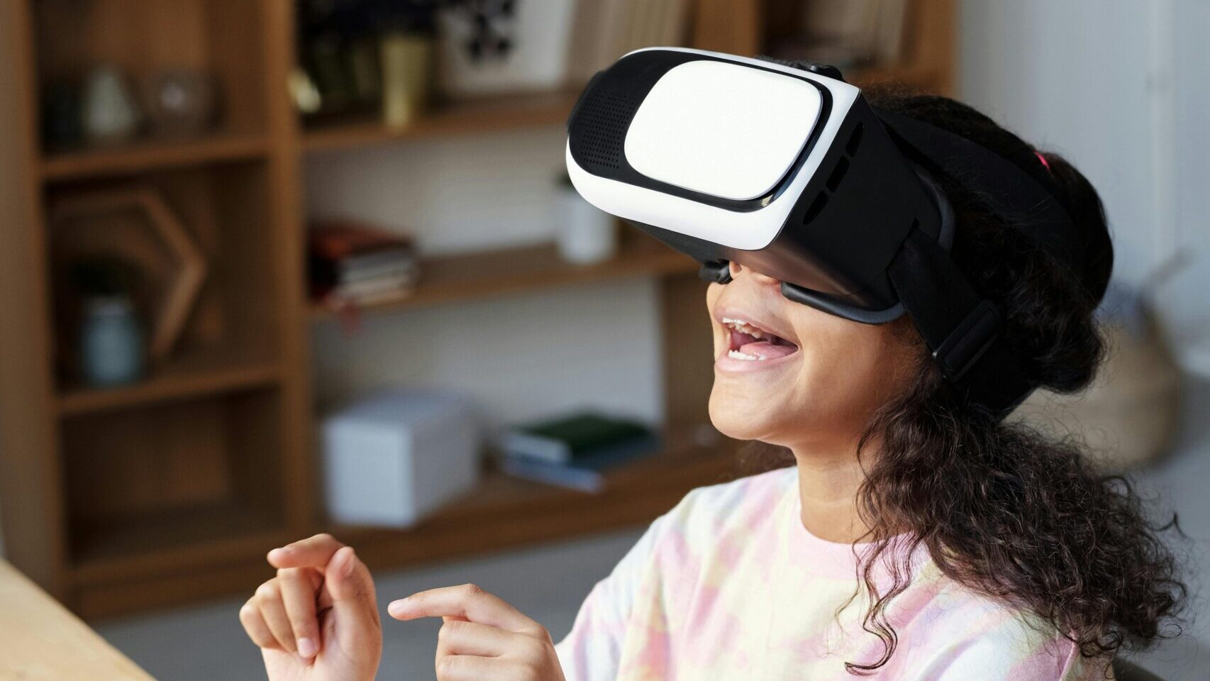 A young girl sitting at a desk while smiling with a VR headset on....