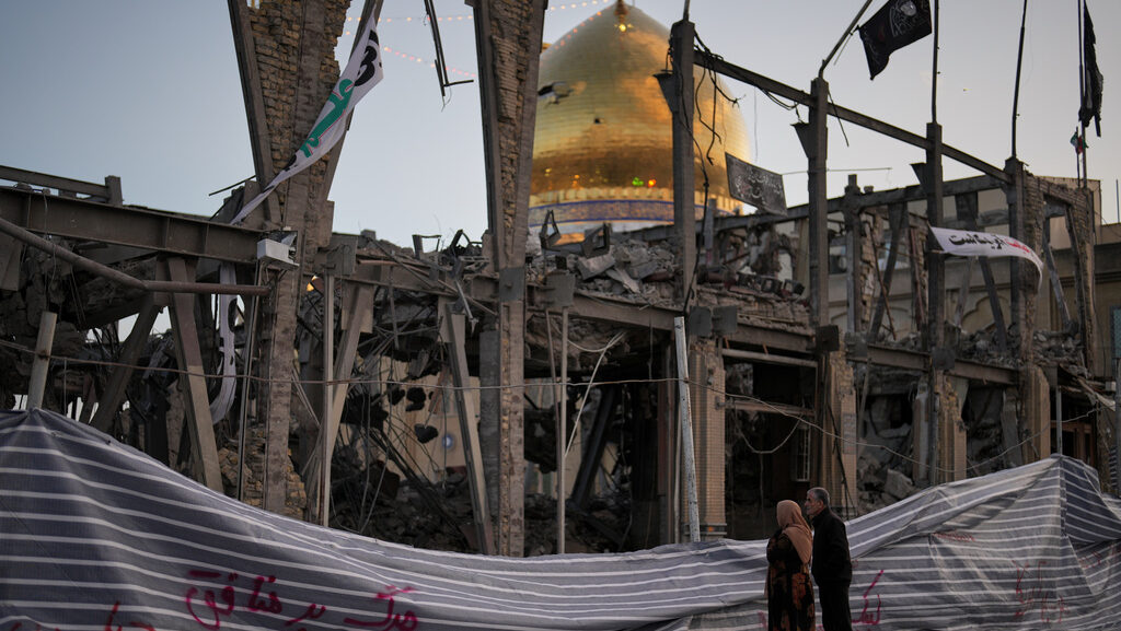 Pedestrians look at a destroyed building within the Grand Hosseiniyeh, with the mosque visible in t...