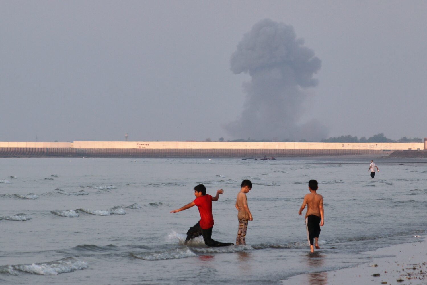 Three boys play in the shallow waters of the Strait of Hormuz, as a plume of smoke rises from an ex...