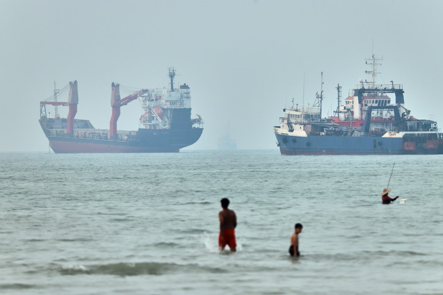 FILE - Children wade in the water with cargo ships at anchor in the background and a fisherman near...
